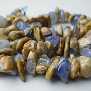 May include: A beaded bracelet composed of polished, irregularly shaped stones in shades of tan, brown, and blue. The stones are strung together with a blue string, creating a natural and earthy aesthetic.