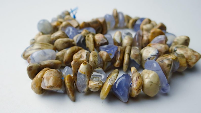 May include: A beaded bracelet composed of polished, irregularly shaped stones in shades of tan, brown, and blue. The stones are strung together with a blue string, creating a natural and earthy aesthetic.
