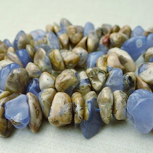 May include: A bracelet made of polished, irregular-shaped stones. The stones are a mix of light blue and tan, creating a natural, earthy aesthetic. The bracelet is strung together with a blue cord.