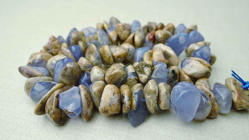 May include: A bracelet made of polished, irregular-shaped stones. The stones are a mix of light blue and tan, creating a natural, earthy aesthetic. The bracelet is strung together with a blue cord.