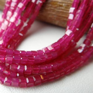 Puede incluir: Primer plano de un collar de varias hebras con cuentas rosas vibrantes, de corte cuadrado. Las cuentas tienen un aspecto facetado, que capta la luz. El collar se muestra sobre un fondo neutro, resaltando el color y la textura.