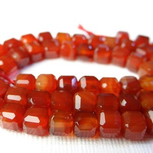 May include: A strand of faceted, reddish-orange gemstone beads. The beads are rectangular with cut corners, strung on a red thread. The gemstones have a polished, reflective surface, and are arranged in a curved shape.