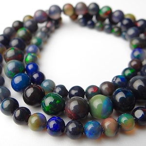 May include: Strands of iridescent opal beads in various sizes and colors, including blue, green, and black. The beads are arranged in a circular pattern, showcasing their polished surfaces.