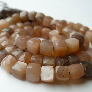 May include: A strand of faceted, square-cut beads in shades of tan, brown, and peach. The beads are strung together, forming a necklace or bracelet. The background is white, highlighting the colors and textures of the gemstones.
