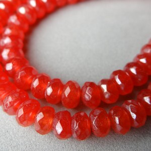 May include: Close-up of two strands of faceted, bright red beads. The beads are small, with a slightly irregular shape, and strung together to form a necklace or bracelet. The beads have a glossy finish.