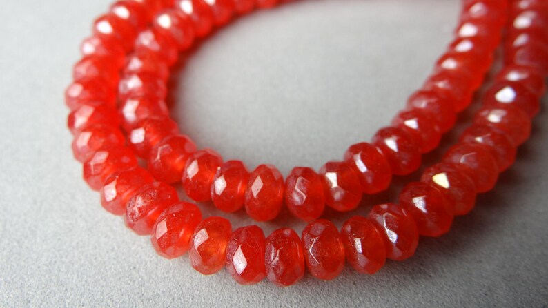 May include: Close-up of two strands of faceted, bright red beads. The beads are small, with a slightly irregular shape, and strung together to form a necklace or bracelet. The beads have a glossy finish.