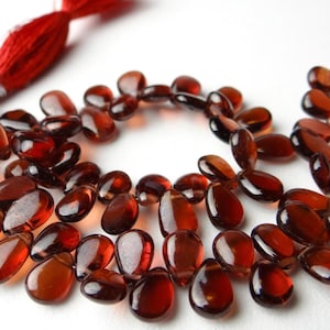 May include: A necklace of polished, teardrop-shaped, dark red gemstones. The beads are strung together, with a red tassel at each end. The gemstones are approximately 1 cm in length.