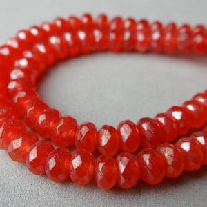 May include: Close-up of a strand of faceted, orange-red gemstone beads. The beads are strung together, forming a necklace or bracelet. The beads have a sparkling, polished appearance, and are arranged in a curved pattern.