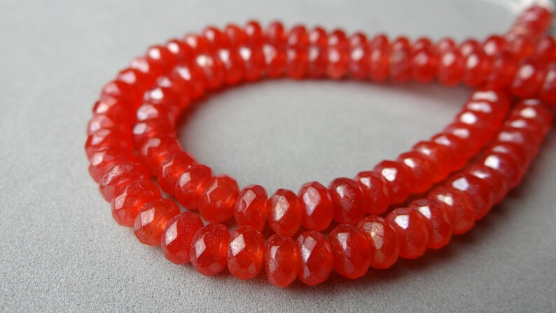 May include: Close-up of a strand of faceted, orange-red gemstone beads. The beads are strung together, forming a necklace or bracelet. The beads have a sparkling, polished appearance, and are arranged in a curved pattern.