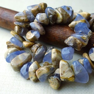 May include: A bracelet made of polished blue and tan gemstones. The stones are irregularly shaped and strung together. The bracelet is resting on a wooden dowel. The gemstones have a translucent quality.