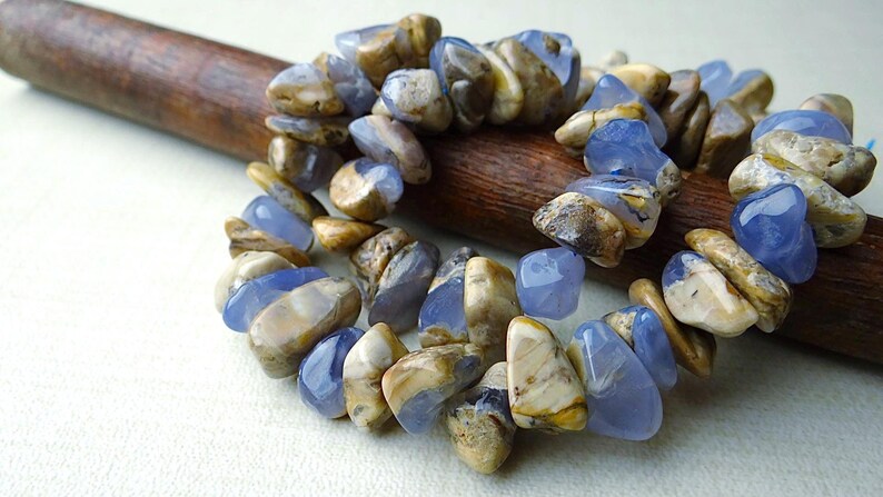 May include: A bracelet made of polished blue and tan gemstones. The stones are irregularly shaped and strung together. The bracelet is resting on a wooden dowel. The gemstones have a translucent quality.