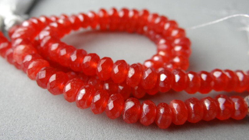 May include: Close-up of a strand of faceted, red gemstone beads. The beads are strung together, forming a necklace or bracelet. The beads have a glossy, transparent appearance, and the color is a vibrant, warm red.