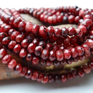 May include: A strand of faceted, round beads in deep shades of red and burgundy with dark, mottled black inclusions. The beads are polished to a high shine, creating a reflective surface that highlights the varied, natural-looking color patterns.
