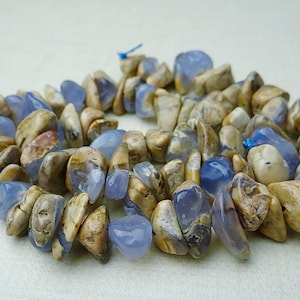 May include: A necklace made of polished, irregular-shaped stones. The stones are a mix of light blue and tan, with a slightly glossy finish. The necklace is strung on a blue cord, and the stones are tightly packed together.