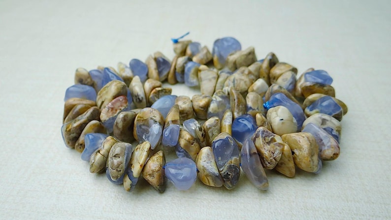 May include: A necklace made of polished, irregular-shaped stones. The stones are a mix of light blue and tan, with a slightly glossy finish. The necklace is strung on a blue cord, and the stones are tightly packed together.