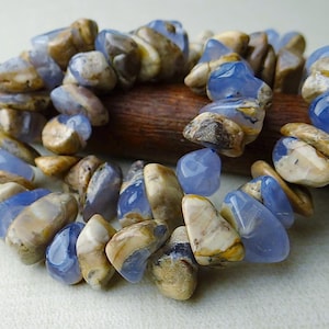 May include: A bracelet made of polished, irregular-shaped blue and tan gemstones. The stones are strung together, creating a flexible band. The bracelet is resting on a wooden dowel against a neutral background.