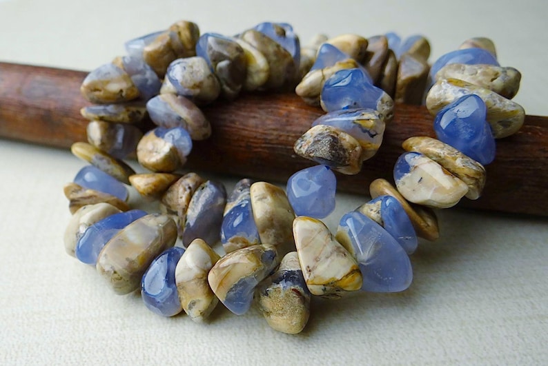 May include: A bracelet made of polished, irregular-shaped blue and tan gemstones. The stones are strung together, creating a flexible band. The bracelet is resting on a wooden dowel against a neutral background.