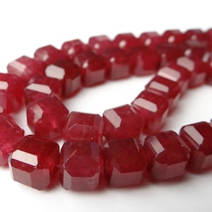 May include: A strand of faceted, deep red beads. The beads are rectangular with multiple cut surfaces, creating a sparkling effect. The beads are strung together, forming a curved shape against a white background. Ideal for jewelry making.