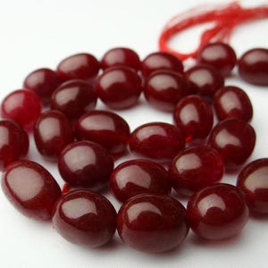 May include: A strand of polished, oval-shaped beads in a deep burgundy color. The beads are strung together with a red cord, creating a circular arrangement against a white background. The beads have a glossy finish.