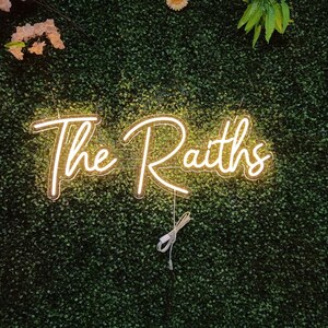 May include: A neon sign with the words "The Raiths" in a cursive font, illuminated in a warm, golden light. The sign is mounted on a backdrop of dense, green foliage, with a few floral accents.
