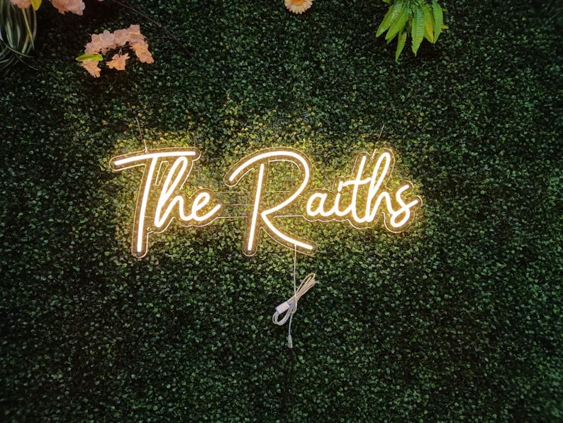 May include: A neon sign with the words "The Raiths" in a cursive font, illuminated in a warm, golden light. The sign is mounted on a backdrop of dense, green foliage, with a few floral accents.