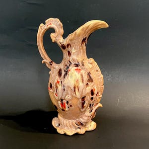 May include: A decorative ceramic pitcher with a tan base, adorned with brown, red, and white accents. The pitcher features an ornate handle and spout, with a textured surface. The base has a decorative, scalloped design.