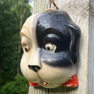 May include: A ceramic wall plaque shaped like a dog head with a glossy finish. The dog has a cream-colored face with a large black patch over the left eye, a black nose, orange accents around the right eye, and bright red markings on the lower chin.