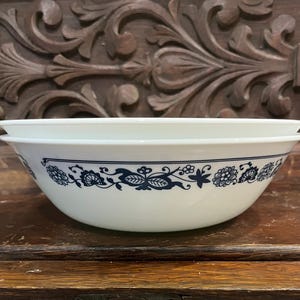 May include: A white glass bowl featuring a dark blue floral pattern around the exterior rim. The bowl has a smooth, rounded shape with a wide opening and a clean, minimalist design. The pattern consists of repeating stylized flowers and leaves.