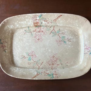 May include: Rectangular serving platter with rounded corners. The beige ceramic platter features a floral design with pink and green blossoms on brown branches. The surface has a speckled pattern.