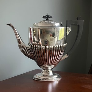 May include: A silver-plated teapot with a black handle and lid finial. The teapot has a fluted lower body and a curved spout. The design is classic and elegant, suitable for serving tea or coffee.
