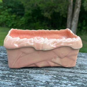 May include: A rectangular ceramic planter in a soft pastel pink glaze. The exterior features a textured, overlapping ribbon or fabric-like design with a ruffled, scalloped top edge. The finish is glossy, reflecting light across the curved surfaces.
