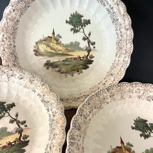 May include: A set of three vintage cream-colored ceramic plates. Each plate features a scalloped edge with gold floral detailing. The center of each plate displays a landscape scene with a church, trees, and figures. The plates are likely for decorative use.