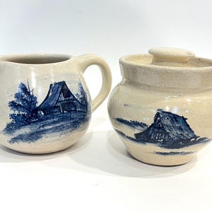 May include: Cream-colored ceramic creamer and sugar bowl set. Each piece features a hand-painted blue barn scene. The creamer has a handle and spout, while the sugar bowl has a lid. The set is ideal for serving coffee or tea.
