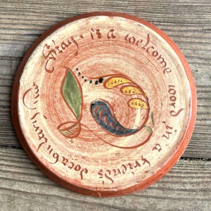 May include: A round terracotta plate featuring a hand-painted design of a green leaf and a blue paisley shape with yellow accents. The text "Stay is a welcome word in a friend's vocabulary" is inscribed in a circular pattern around the center.