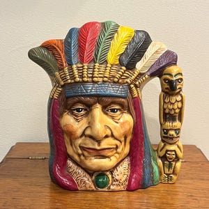 May include: A ceramic mug featuring a Native American chief design. The mug is painted with vibrant colors, including red, yellow, blue, and green. It has a handle with a totem pole design and a decorative feather headdress.