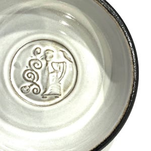 May include: A handmade ceramic bowl with a white interior and a dark rim. The bowl features a raised relief design of a figure with wings and swirling patterns in the center. The bowl is a unique piece of pottery.
