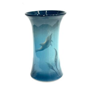 May include: A blue ceramic vase with a dolphin design. The vase has a flared top and a cylindrical body. The dolphin is depicted swimming in a blue ocean with seaweed. The vase is approximately 8 inches tall.