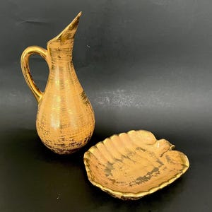 May include: A gold-toned ceramic pitcher with a textured surface and a curved handle. Beside it is a leaf-shaped dish, also gold-toned. Both items have a metallic finish and are set against a dark background.
