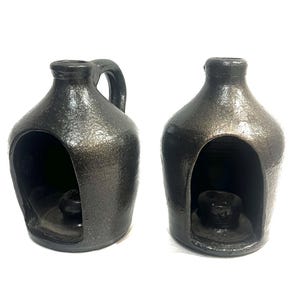 May include: Two dark gray ceramic candle holders. One has a handle, and both have a large opening in the front. Each candle holder has a small, round base inside to hold a candle. The candle holders have a textured surface.