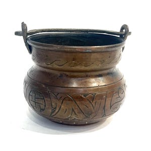 May include: A small, antique copper pot with a handle. The pot has decorative etched patterns around the body and a dark patina. The handle is attached to the rim with metal loops.