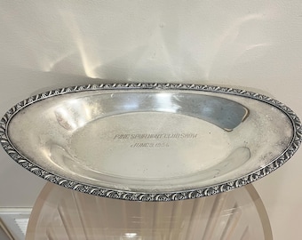 May include: An oval silver-toned serving tray with a decorative embossed border. The center of the tray features an engraving that reads: PINE SPUR HUNT CLUB SHOW JUNE 9, 1956. The surface has a polished finish with visible signs of age and patina.