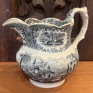 May include: A cream-colored ceramic pitcher featuring a black transferware pattern of an orientalist landscape with domed buildings, palm trees, and mountains. The pitcher has a scalloped rim, a curved handle, and a detailed floral border design.