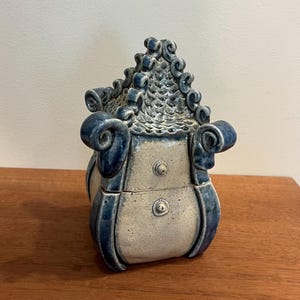 May include: A ceramic lidded jar with a light beige speckled body and a dark blue glazed roof. The roof features a textured scale pattern and curled edges. The jar has a curved silhouette with two small circular protrusions on the front panel.