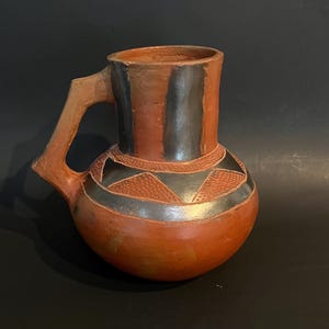 May include: A handmade terracotta pitcher with a handle. The pitcher has a rounded base and a cylindrical neck. It features a reddish-brown body with black and gray geometric designs around the neck and vertical stripes. The handle is a lighter shade of brown.