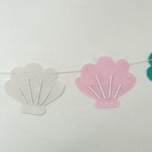 May include: A seashell garland made of pink, teal, and white felt cutouts. The seashell shapes are strung on a white string. The banner is designed for decoration and is set against a plain white backdrop.