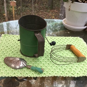 以下が含まれることがあります： 緑色の金属製の小麦粉ふるい、銀色のさじ、木製のハンドルが付いた生地ミキサーが緑色のランチョンマットの上に置かれています