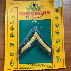 May include: A package of three pairs of green and gold chevron patches. The package is yellow with a green background and the text "CHEVRONS WITH AUTHENTICITY" in white. The text "HALLMARK A 21" is also printed on the package.
