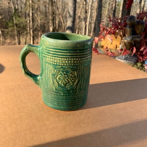 Pode incluir: Uma caneca de cerâmica verde com um desenho em relevo de um cacho de uvas. A caneca tem uma alça e é decorada com uma faixa de pontos brancos ao redor da parte superior.