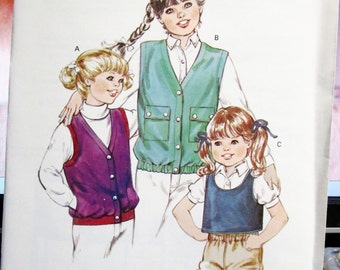 May include: A sewing pattern for three children's vests. The pattern is called "Kwik-Sew" and is for sizes 8-10-12-14. The pattern shows three girls wearing different colored vests. The vest on the left is purple, the vest in the middle is green, and the vest on the right is blue.