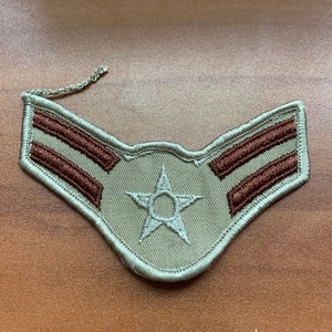Puede incluir: Un parche de rango militar de color beige con dos rayas marrones y una estrella blanca con un círculo en el centro.